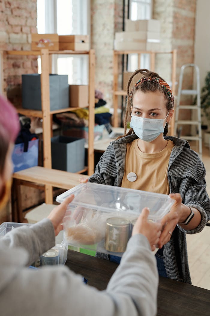 A young woman in a mask hands a food donation box in an indoor facility.